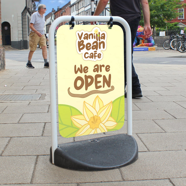 Outdoor signage for businesses in the UK featuring an EcoSwinger sign with light yellow background, showcasing 'Vanilla Bean Café' and the text 'We are OPEN.' Includes a vanilla flower illustration, perfect for attracting customer attention and enhancing visibility.