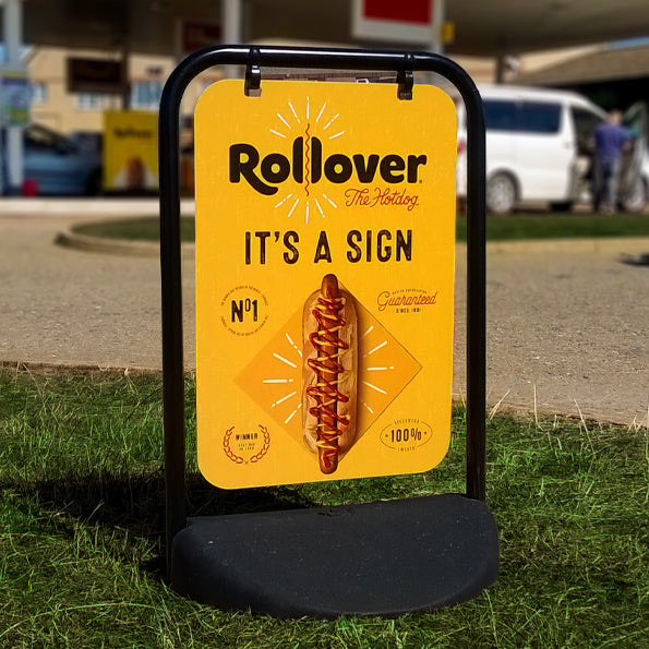 Outdoor signage for businesses in the UK featuring the EcoSwinger sign with bright yellow panel, showcasing the text 'Rollover The Hotdog' along with bold illustrations of a hotdog. Ideal for enhancing visibility and attracting attention in outdoor settings.