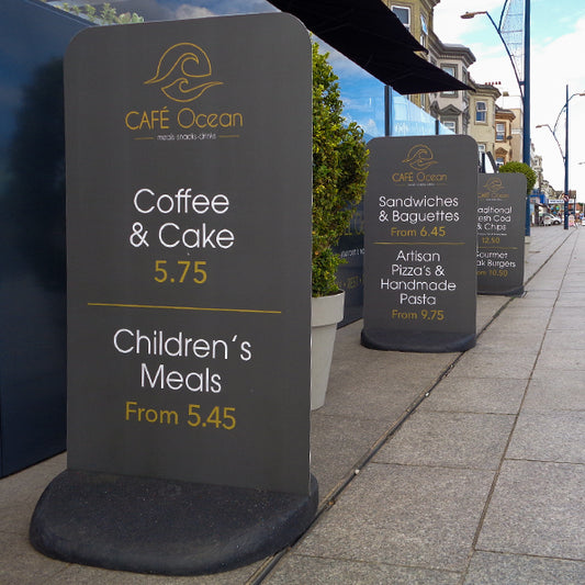 Three Flex Pavement sign boards outside Café Ocean, advertising menu items like coffee, cake, sandwiches, pizzas, cod and chips, and steak burgers with their respective prices. Ideal for outdoor café signage in the UK.